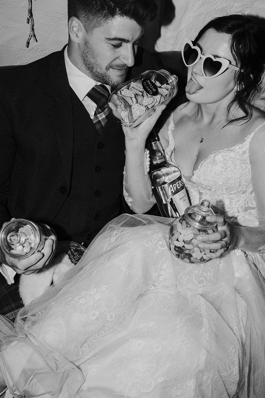 Fun black and white wedding photo of bride and groom sharing Aperol and retro sweet jars at their evening reception
