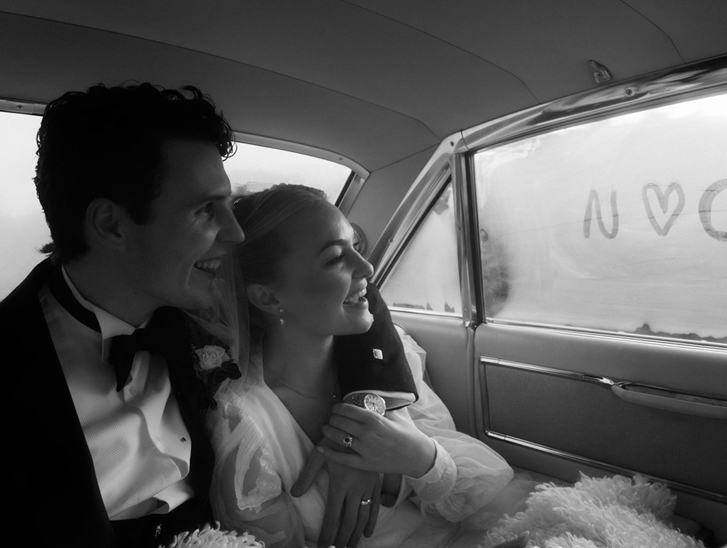 Black and white photo of newly married couple laughing together inside their wedding car with “N loves C” written on the window