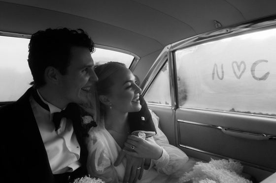 Black and white photo of newly married couple laughing together inside their wedding car with “N loves C” written on the window