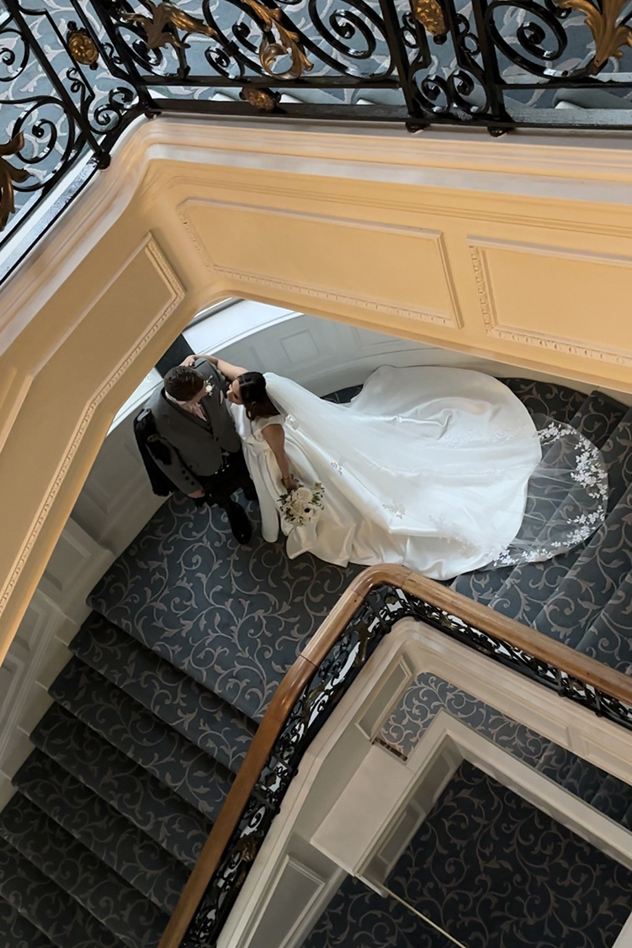 Bride and groom walking down grand staircase together, veil flowing behind in elegant hotel wedding setting