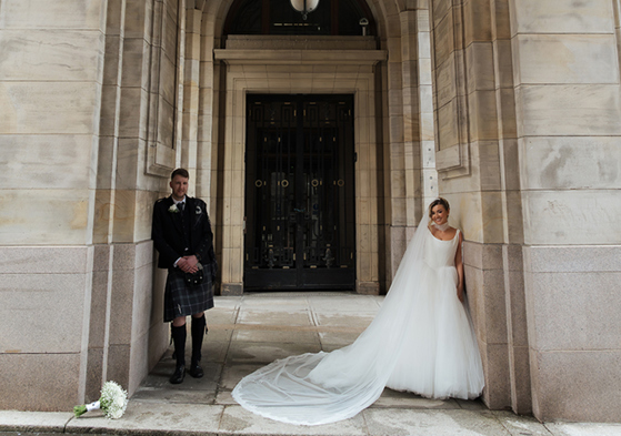 Bride and groom posing under grand stone archway with long veil at elegant city wedding venue
