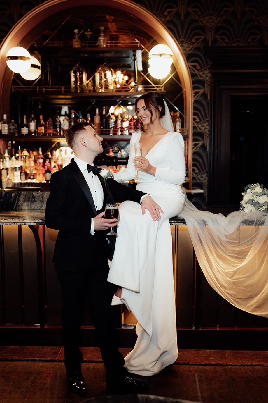 Bride in modern wedding dress sitting on bar with groom in tuxedo holding drinks at stylish city wedding venue