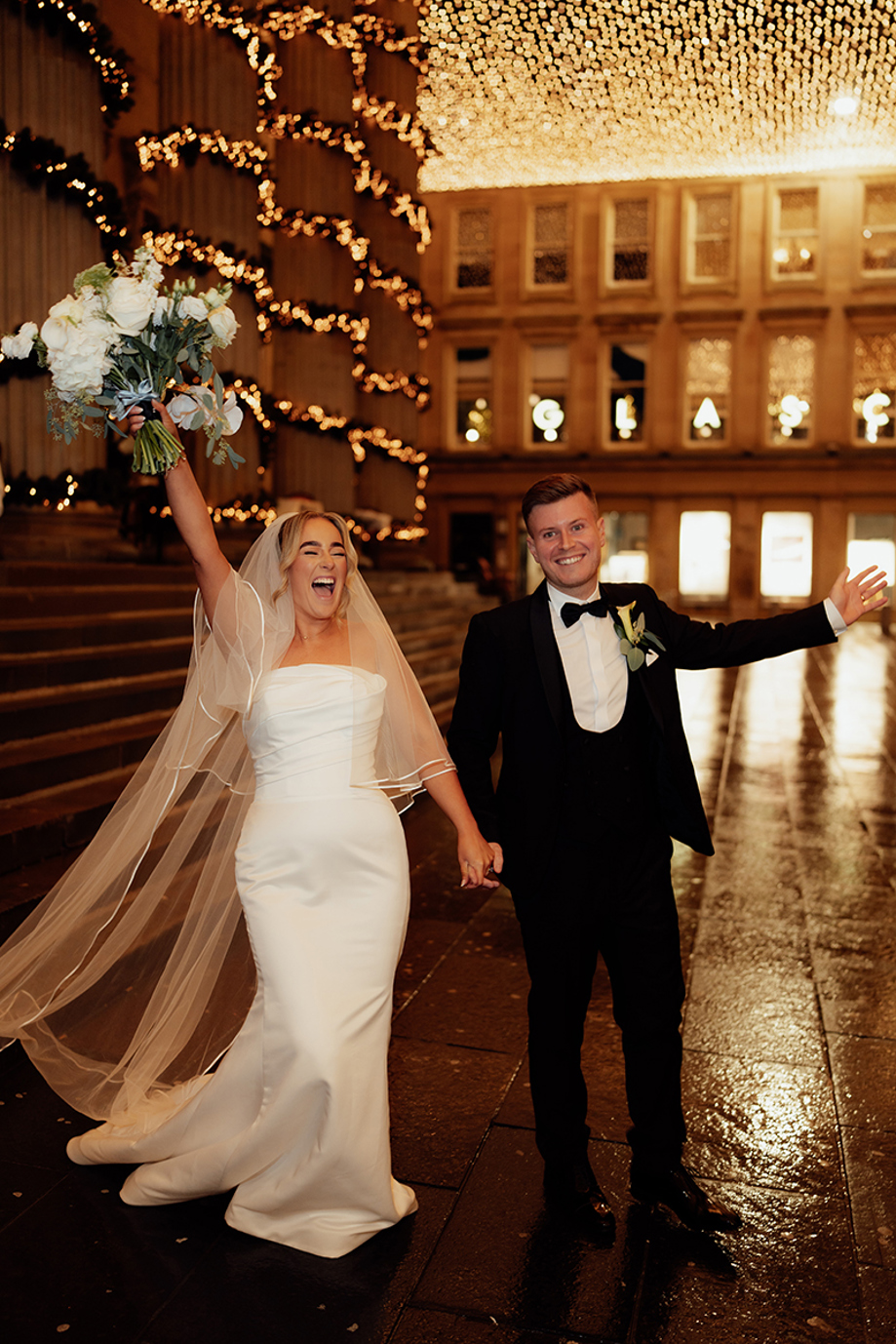 Bride and groom celebrating at night under city lights and fairy lights after wedding ceremony