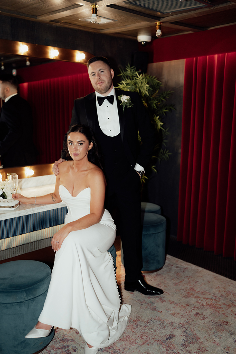 Bride seated in fitted wedding dress with groom standing behind in red velvet lounge editorial wedding portrait