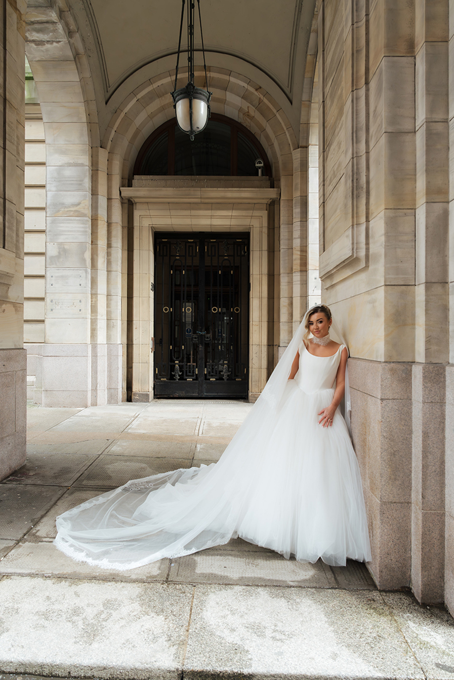 Bride in full skirt wedding dress with long veil leaning against stone archway at city venue