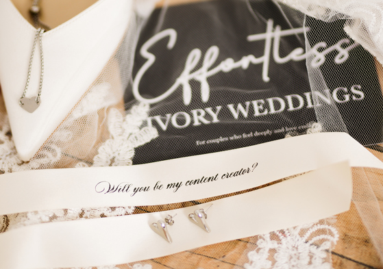 Bridal detail flat lay featuring jewellery, lace wedding accessories and a personalised “Will you be my content creator?” ribbon styled by Effortless Ivory Weddings