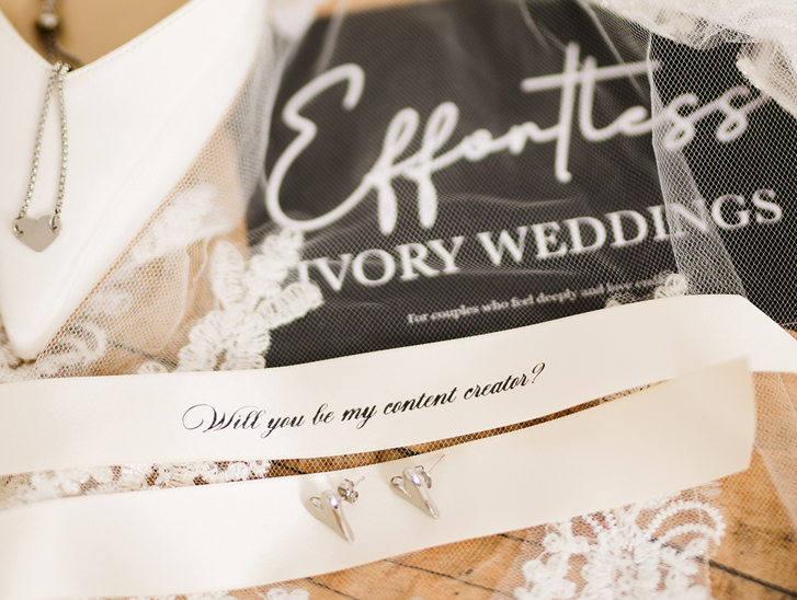 Bridal detail flat lay featuring jewellery, lace wedding accessories and a personalised “Will you be my content creator?” ribbon styled by Effortless Ivory Weddings