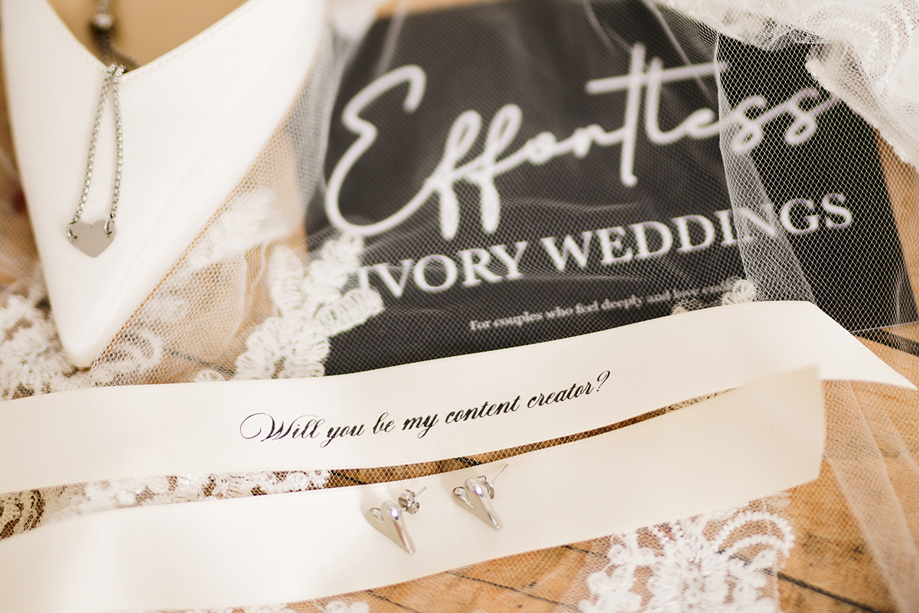 Bridal detail flat lay featuring jewellery, lace wedding accessories and a personalised “Will you be my content creator?” ribbon styled by Effortless Ivory Weddings