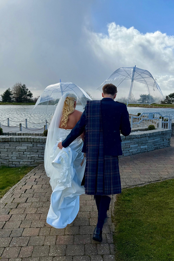 Bride and groom walking beside a lakeside wedding venue holding clear umbrellas on a windy day captured by Arauthentic Moments