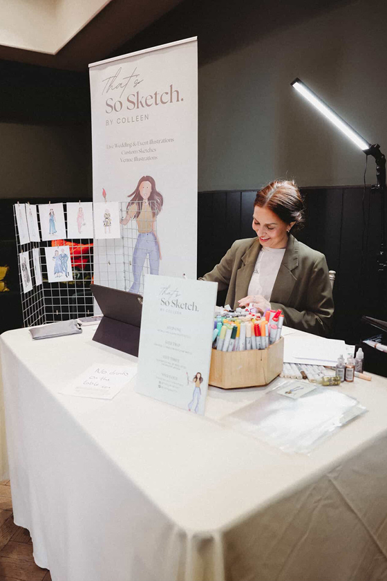 Colleen from That’s So Sketch creating live wedding illustrations at her event setup surrounded by pens and guest sketches.