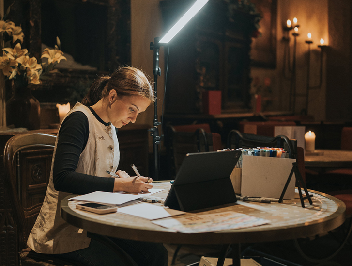 Colleen creating live wedding illustrations during an evening reception with drawing materials and lighting setup.