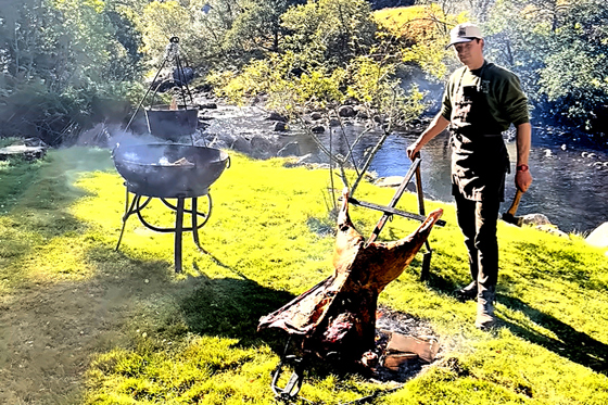 Wylie & Wood chef cooking whole meat over an open fire beside a Scottish river in a Highland setting.