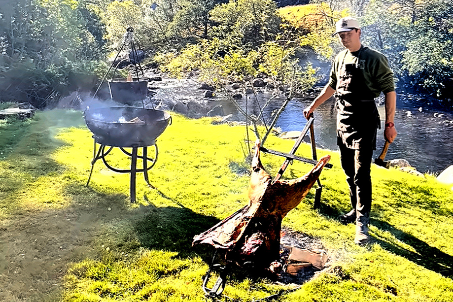 Wylie & Wood chef cooking whole meat over an open fire beside a Scottish river in a Highland setting.