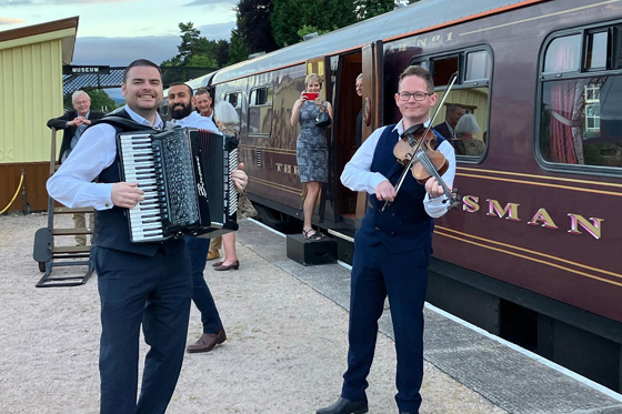 Accordion player and fiddler performing outdoors beside a train for wedding guests.