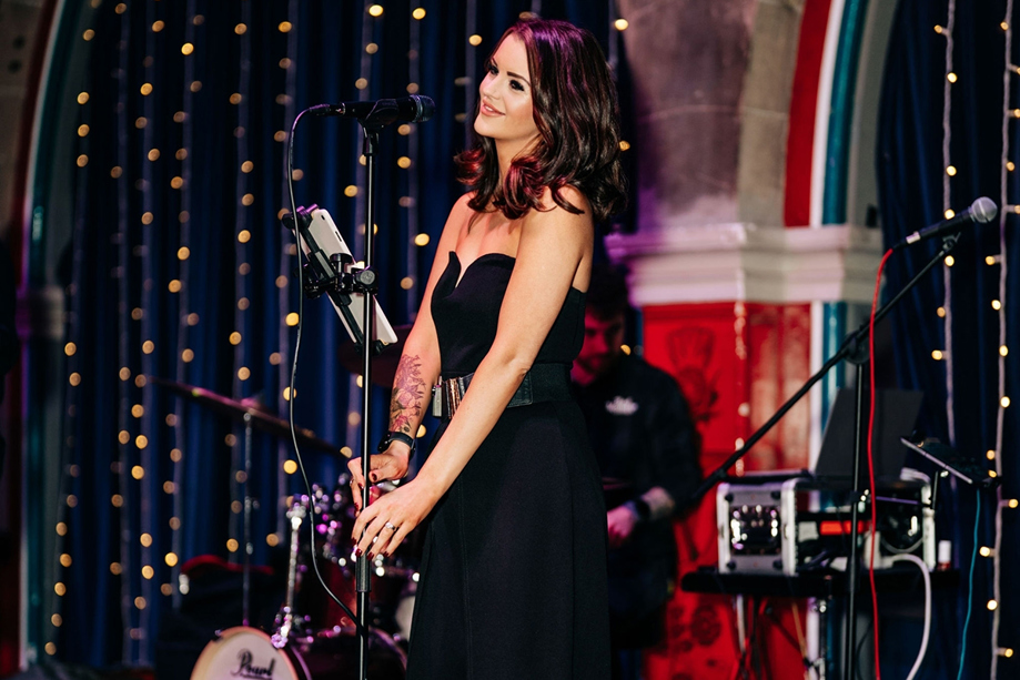 Vocals by Suzie performing live at a wedding or event with microphone, band setup and fairy lights