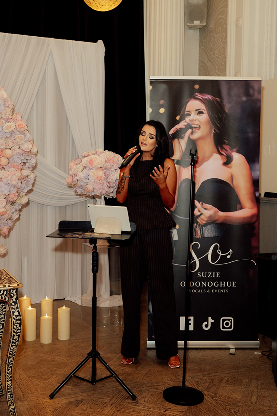 Vocals by Suzie singing at a wedding showcase with branded banner, pink floral arch and candle décor