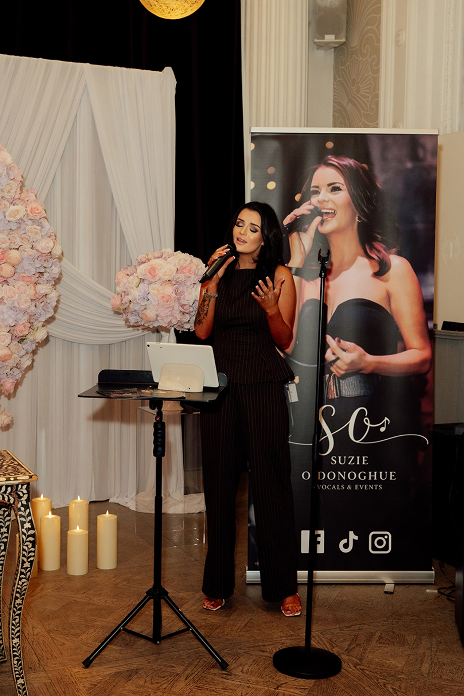 Vocals by Suzie singing at a wedding showcase with branded banner, pink floral arch and candle décor