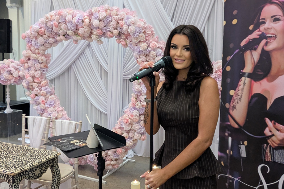 Vocals by Suzie at a wedding showcase with microphone, pink floral arch and branded display banner