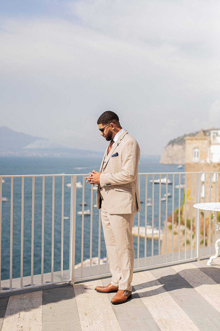 Groom in a beige wedding suit stands on a coastal terrace overlooking the sea before the ceremony.