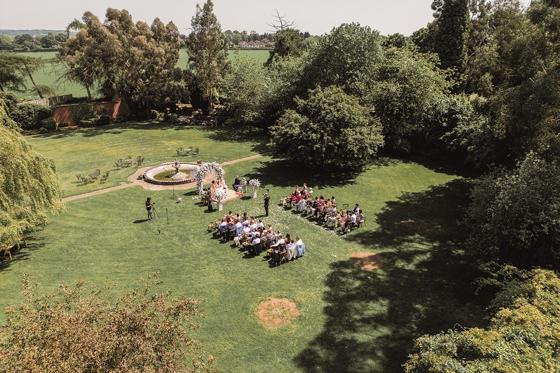 Aerial view of an outdoor wedding ceremony taking place on a large lawn surrounded by mature trees.