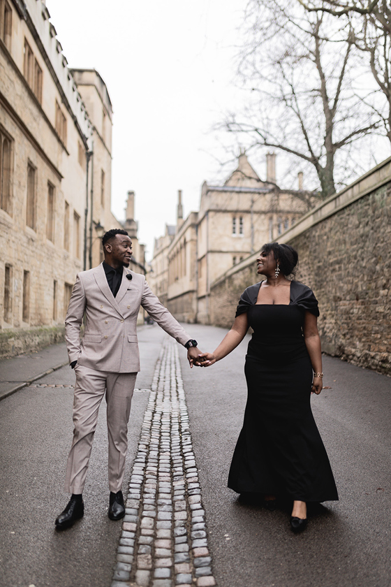 Couple wearing formal evening outfits walk hand in hand along a historic cobbled city street.