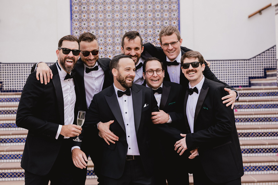 Groom and groomsmen laugh together in black tuxedos during a destination wedding group portrait.