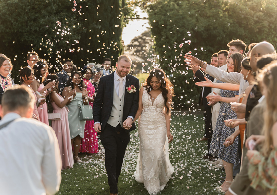 Newlyweds walk hand in hand through a shower of confetti as wedding guests celebrate outdoors.