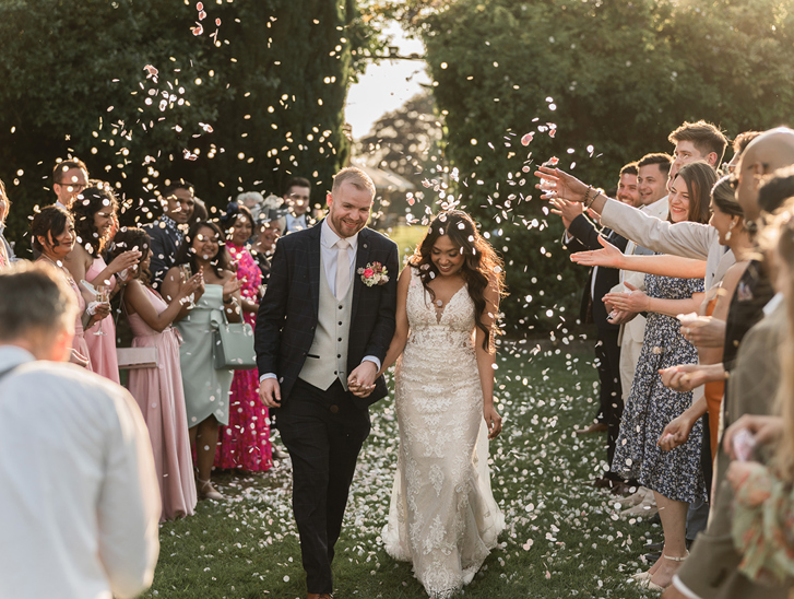 Newlyweds walk hand in hand through a shower of confetti as wedding guests celebrate outdoors.
