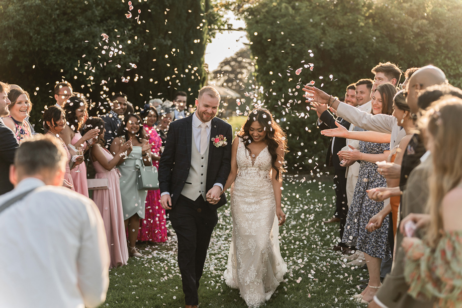 Newlyweds walk hand in hand through a shower of confetti as wedding guests celebrate outdoors.