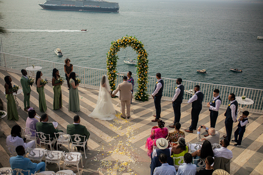 Bride and groom exchange vows beneath a yellow floral arch during an outdoor seaside destination wedding ceremony.