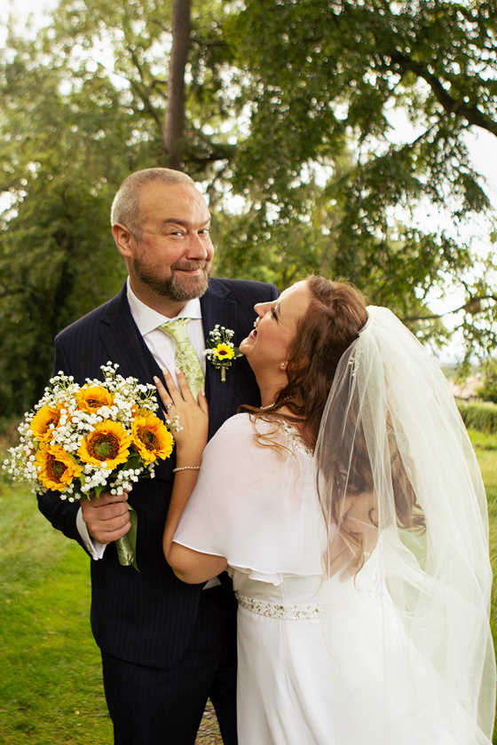 Newlywed bride and groom laughing together outdoors with a bright sunflower wedding bouquet, photographed by Rscott_shoots.