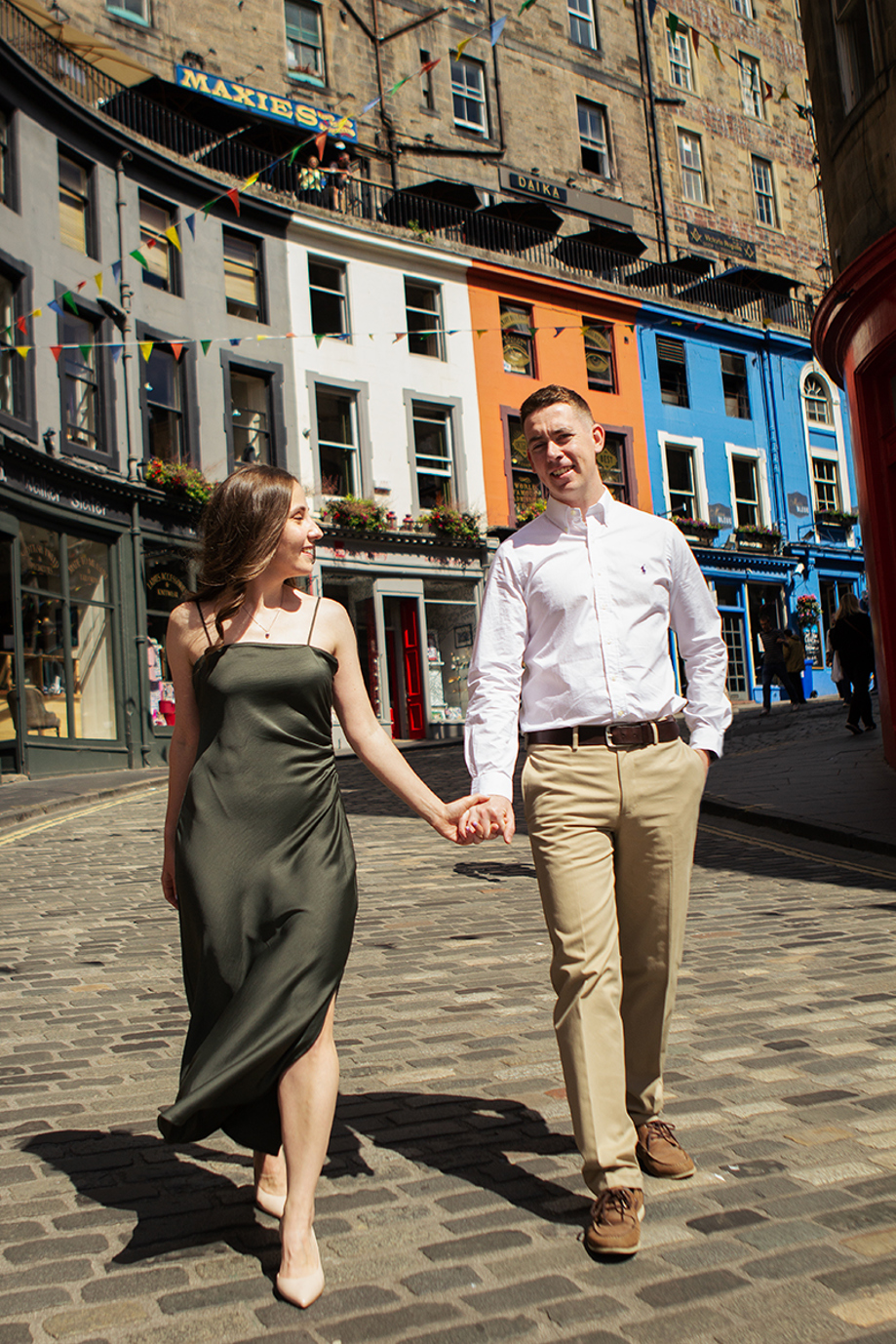 Engaged couple walking hand in hand along colourful Victoria Street in Edinburgh during a city engagement shoot by Rscott_shoots.