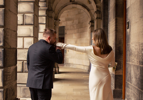 Groom kissing the bride’s hand beneath the stone archways of Edinburgh City Chambers during their city wedding photoshoot by Rscott_shoots.