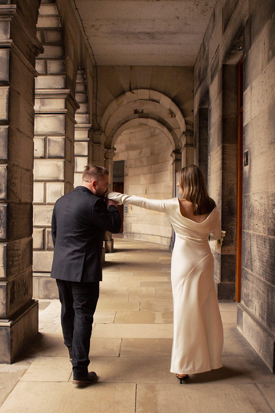 Groom kissing the bride’s hand beneath the stone archways of Edinburgh City Chambers during their city wedding photoshoot by Rscott_shoots.