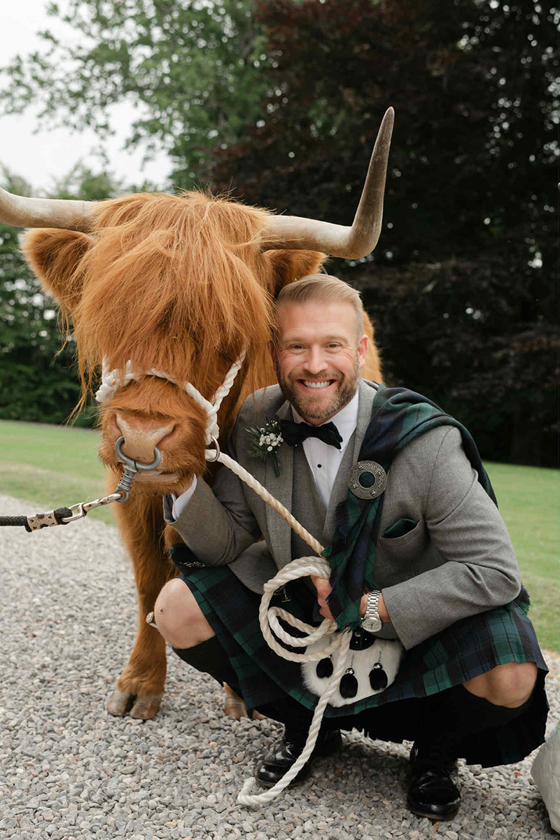 Groom wearing a tartan kilt poses beside a Crazy Critters Experience Highland cow on his wedding day.
