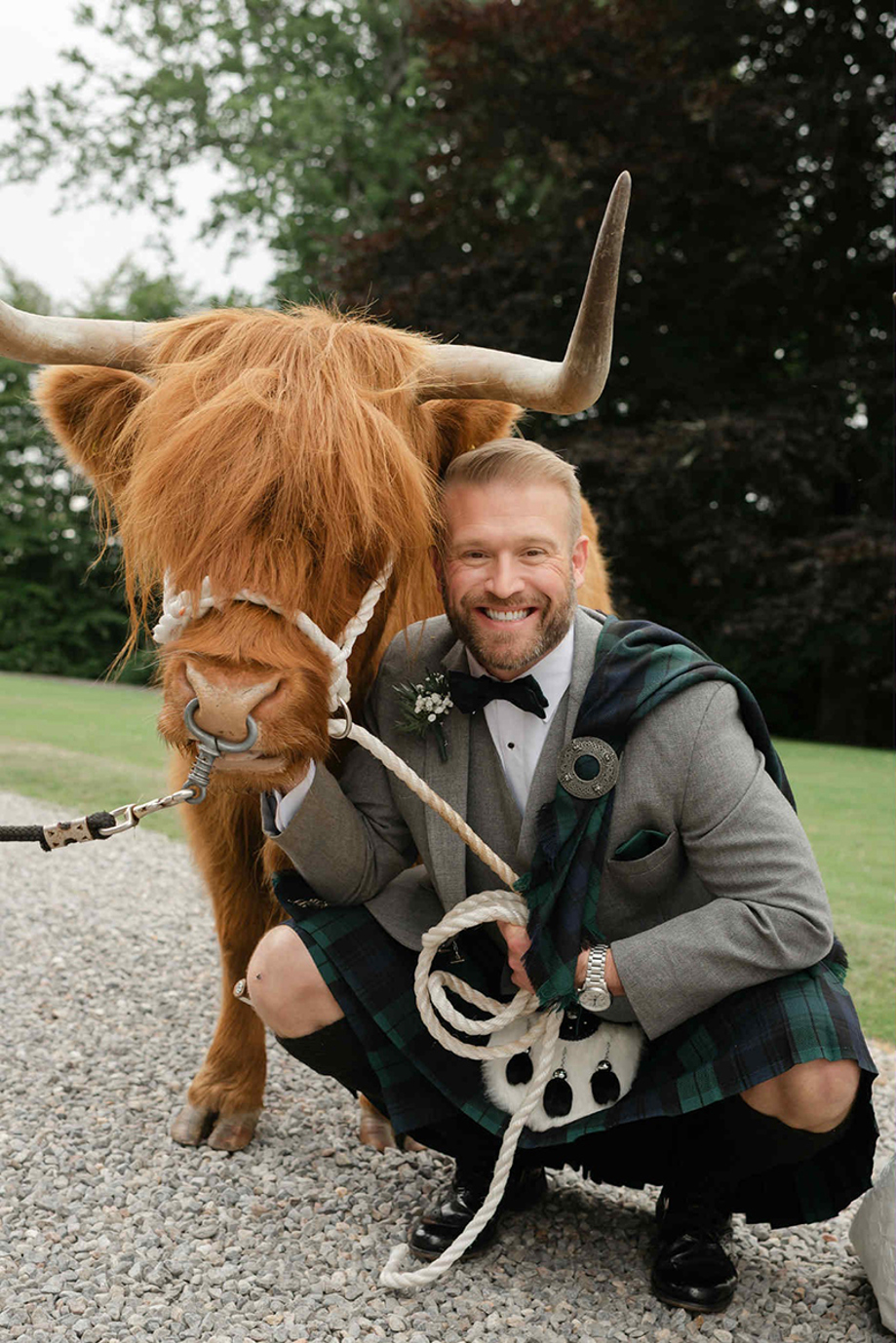 Groom wearing a tartan kilt poses beside a Crazy Critters Experience Highland cow on his wedding day.