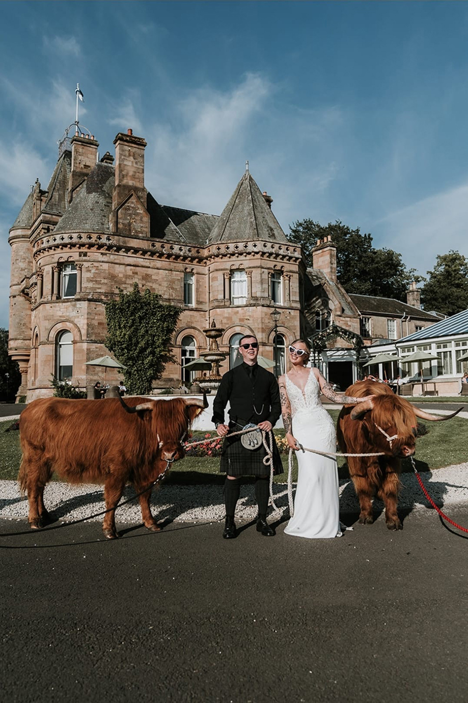 Bride and groom wearing sunglasses pose with two Crazy Critters Experience Highland cows outside a country house wedding venue.