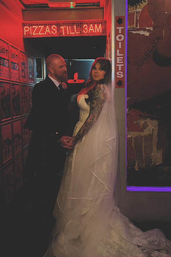 Tattooed bride and groom pose beneath a red neon sign inside a quirky pizza restaurant on their wedding day.