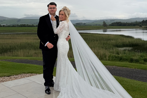 Bride and groom pose outside beside a scenic loch, with the bride’s long veil trailing across the terrace.