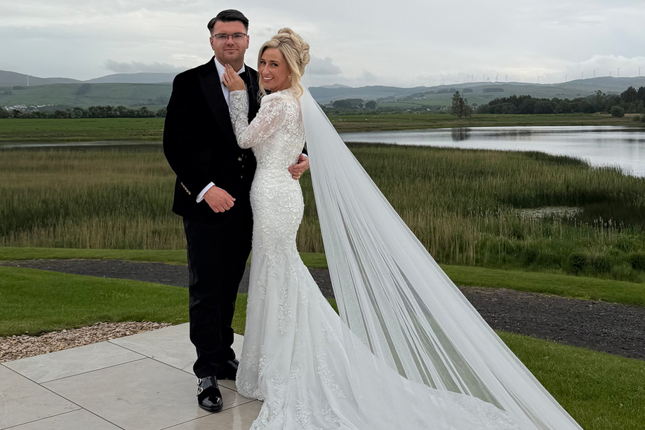 Bride and groom pose outside beside a scenic loch, with the bride’s long veil trailing across the terrace.