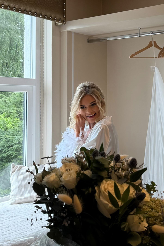 Smiling bride gets ready beside a window with her white and green wedding bouquet in the foreground.