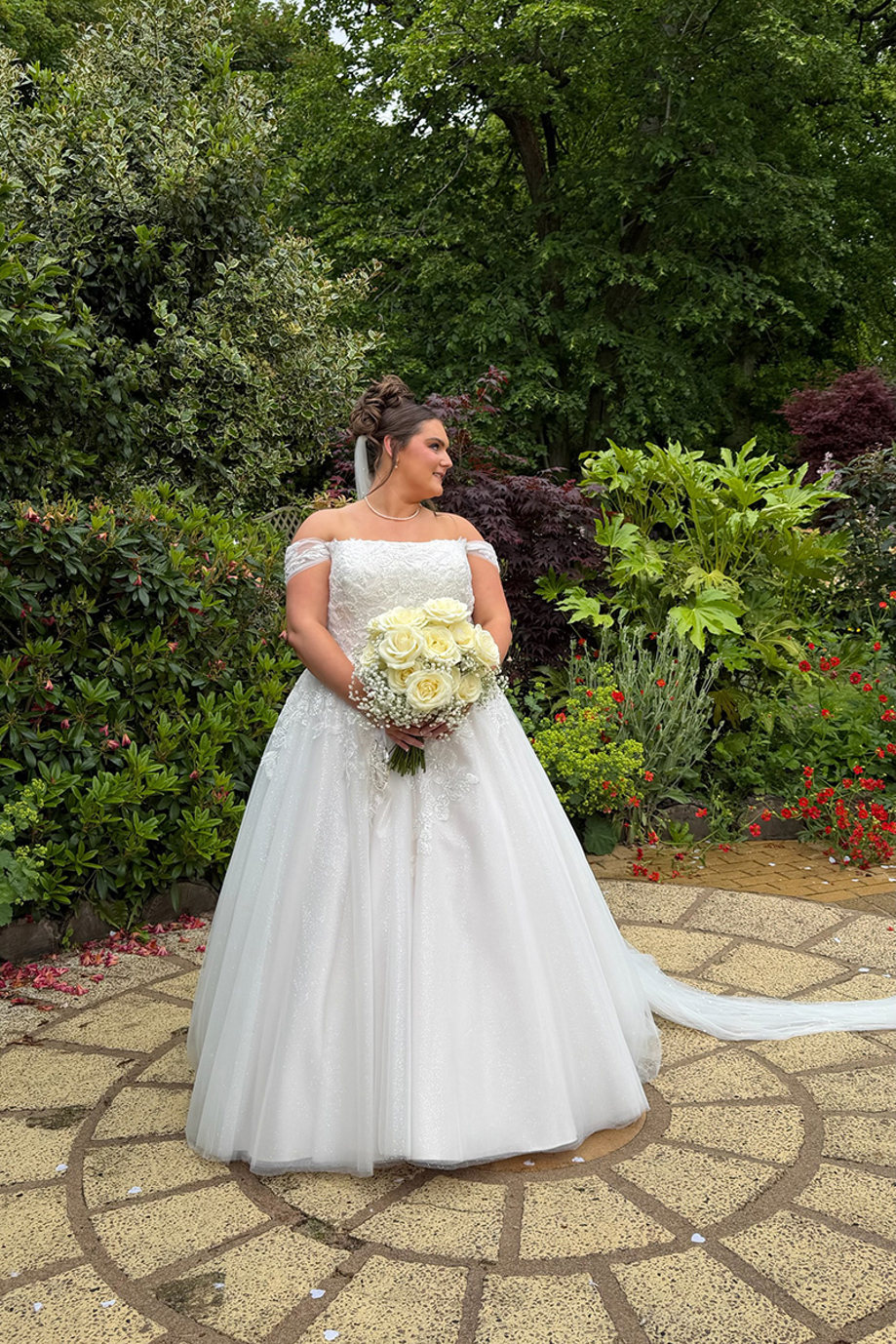 Bride poses in a landscaped wedding venue garden holding a bouquet of white roses and gypsophila.