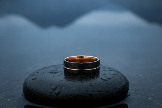 Dark Forge & Lumber Foundry wedding band with copper detailing displayed on a rain-covered stone overlooking a Scottish loch.