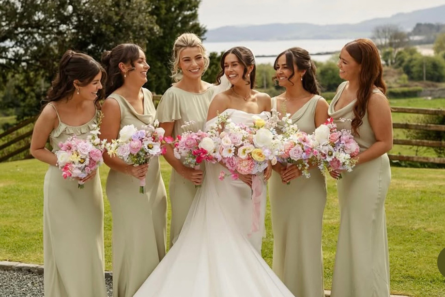 Bride and bridesmaids wearing sage green dresses pose outdoors with colourful wedding bouquets beside a Scottish loch.