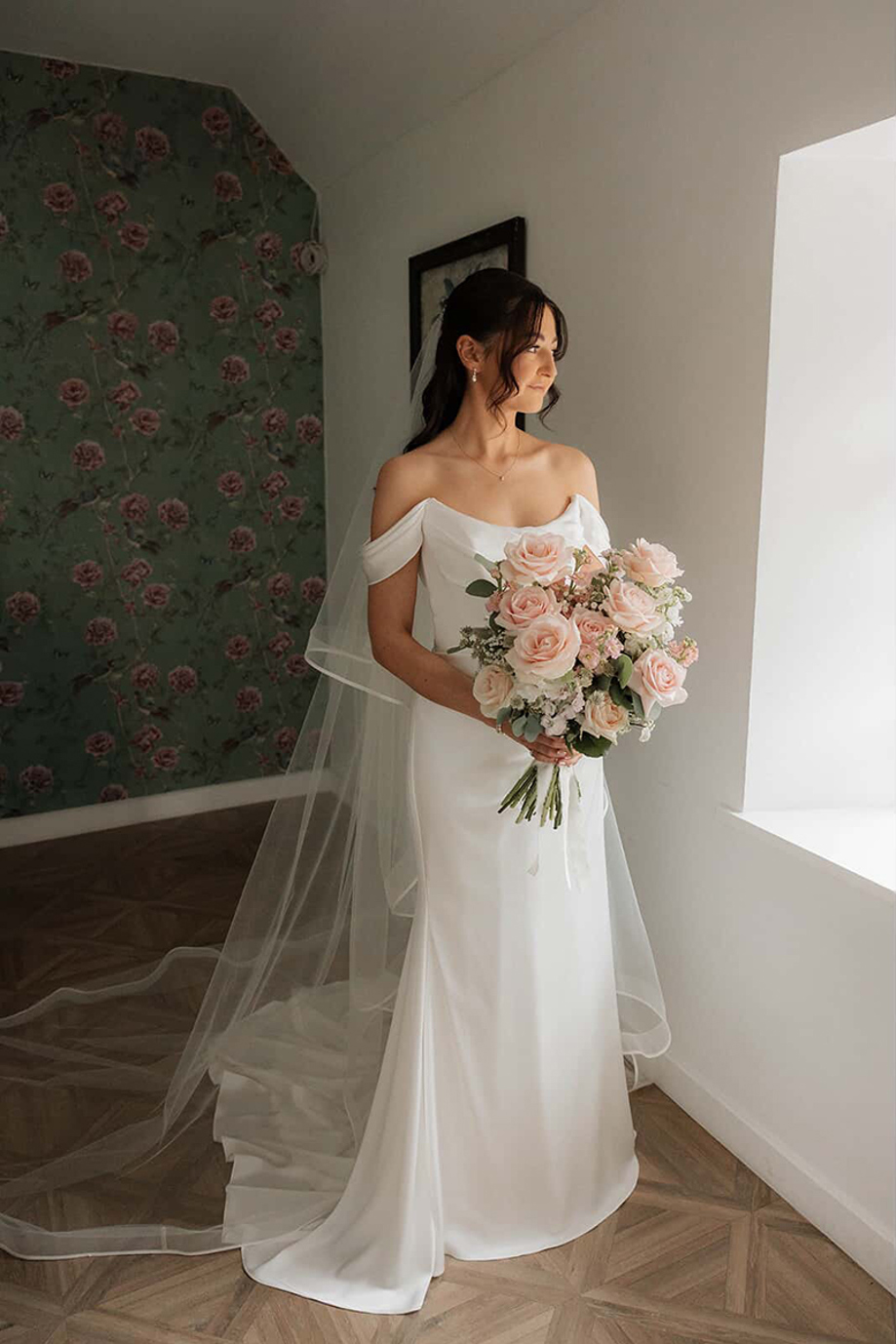 Bride with softly styled brows and lashes poses beside a window holding a bouquet of pale pink roses.