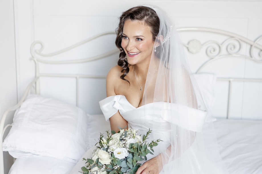 Smiling bride wearing natural wedding beauty styling sits on a bed holding a white and green bridal bouquet.