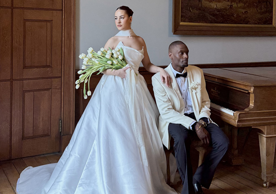Bride holding white tulips stands beside the groom in a cream tuxedo jacket next to a grand piano.