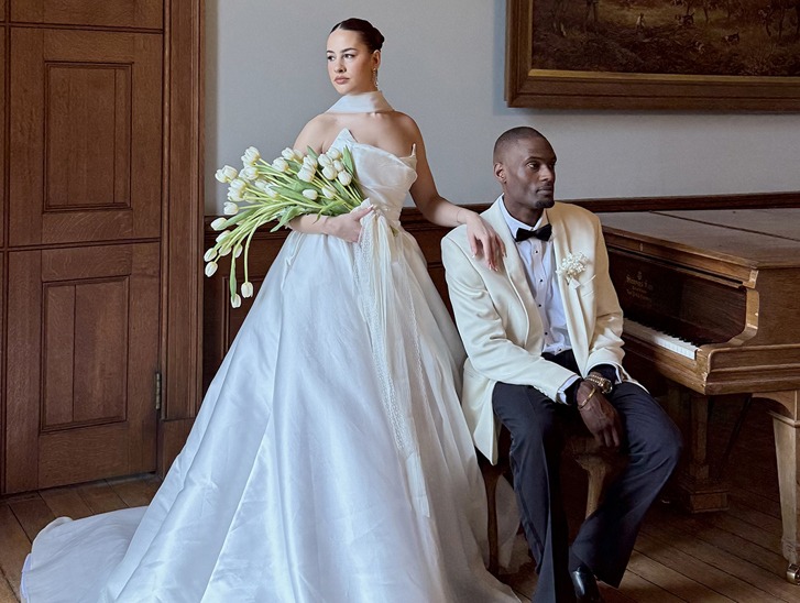 Bride holding white tulips stands beside the groom in a cream tuxedo jacket next to a grand piano. Luxury Wedding Content.