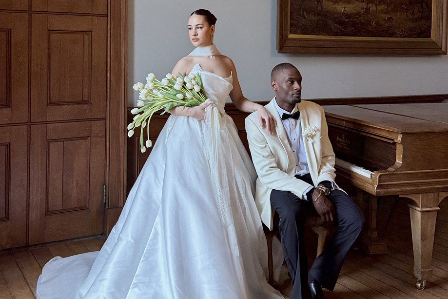 Bride holding white tulips stands beside the groom in a cream tuxedo jacket next to a grand piano.