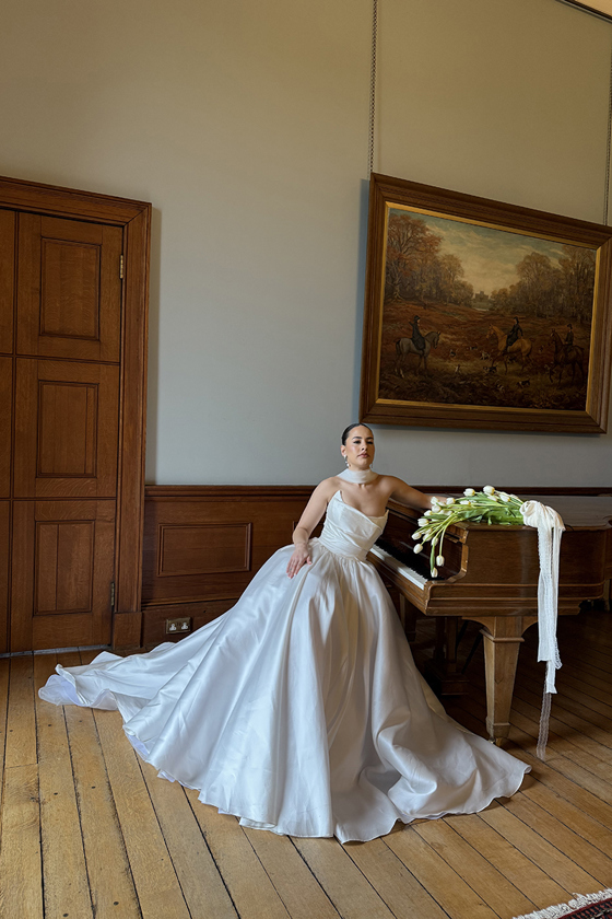 Bride in a full satin wedding dress poses beside a grand piano with a bouquet of white tulips at a historic Scottish venue.